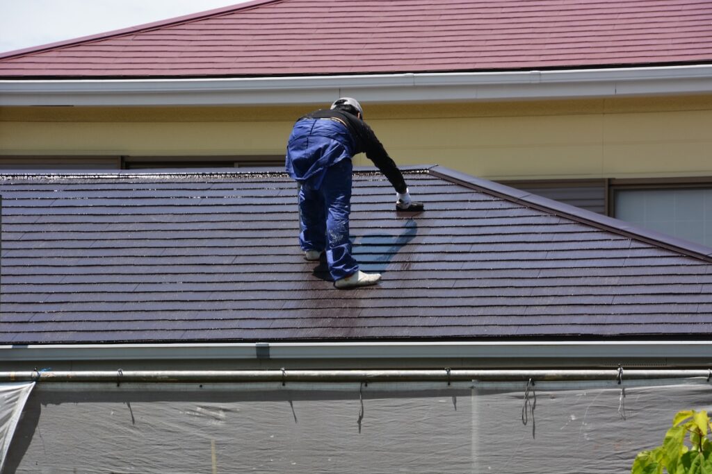 A person wearing protective gear and blue clothing is kneeling on a sloped house roof, performing soft wash roof cleaning. Using a long tool, they clean the wet, shiny shingles with care. soft wash roof cleaning