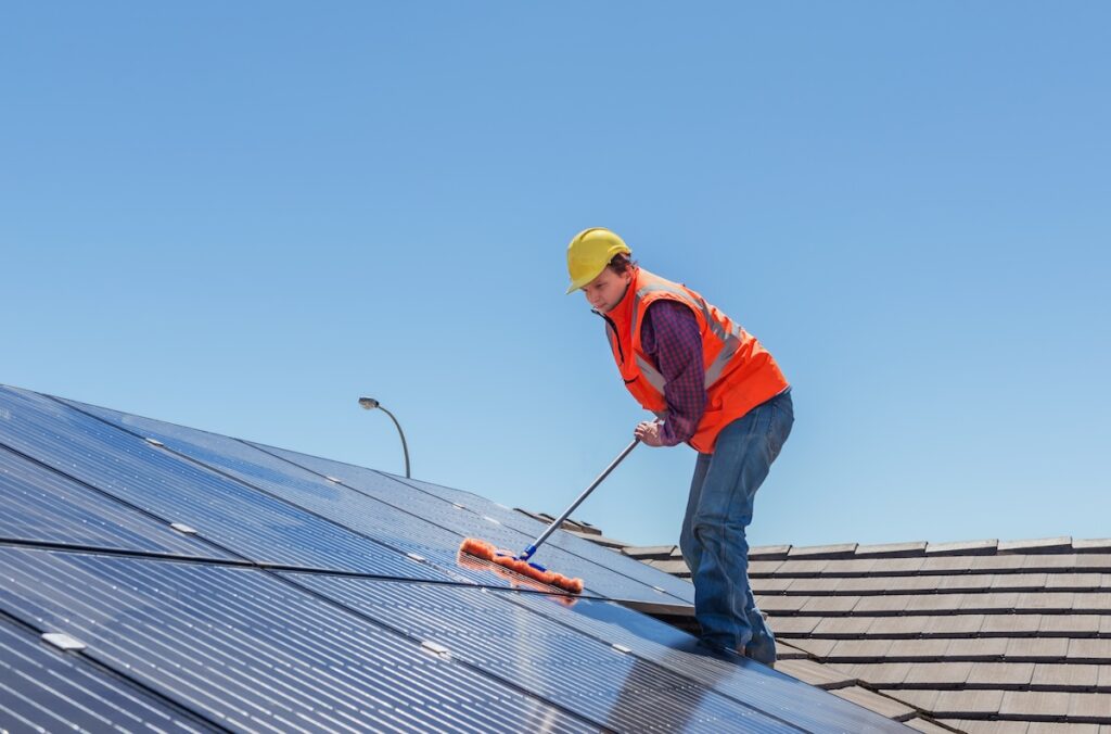 A worker in a yellow hard hat and orange safety vest performs soft wash roof cleaning, using a long-handled brush to clean rooftop solar panels on a sunny day under a clear blue sky. soft wash roof cleaning