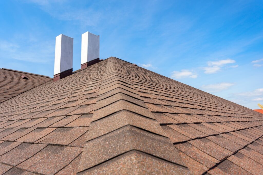 how long do asphalt shingles last Close-up view of a sloped brown shingle roof with two white rectangular chimneys, set against a blue sky with a few scattered clouds.