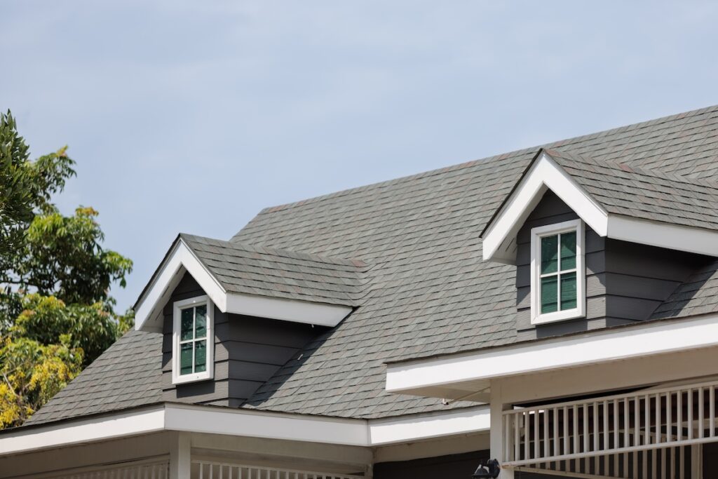 how long do asphalt shingles last The image shows the roof of a house with gray shingles, two dormer windows, and white trim under a partly cloudy sky. Some green tree branches are visible on the left side of the frame.