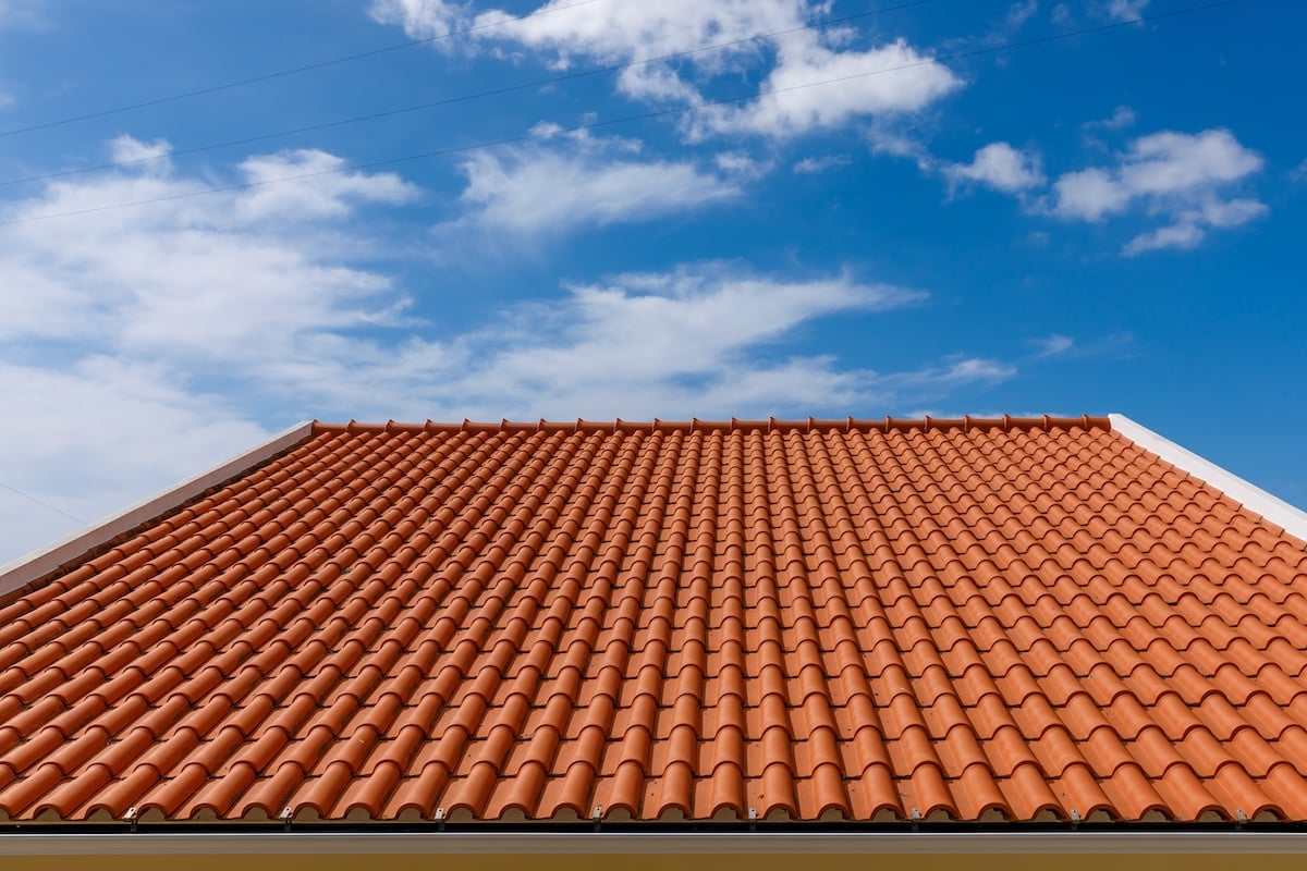 A close-up view of a house roof with orange ceramic tiles under a bright blue sky with scattered white clouds.