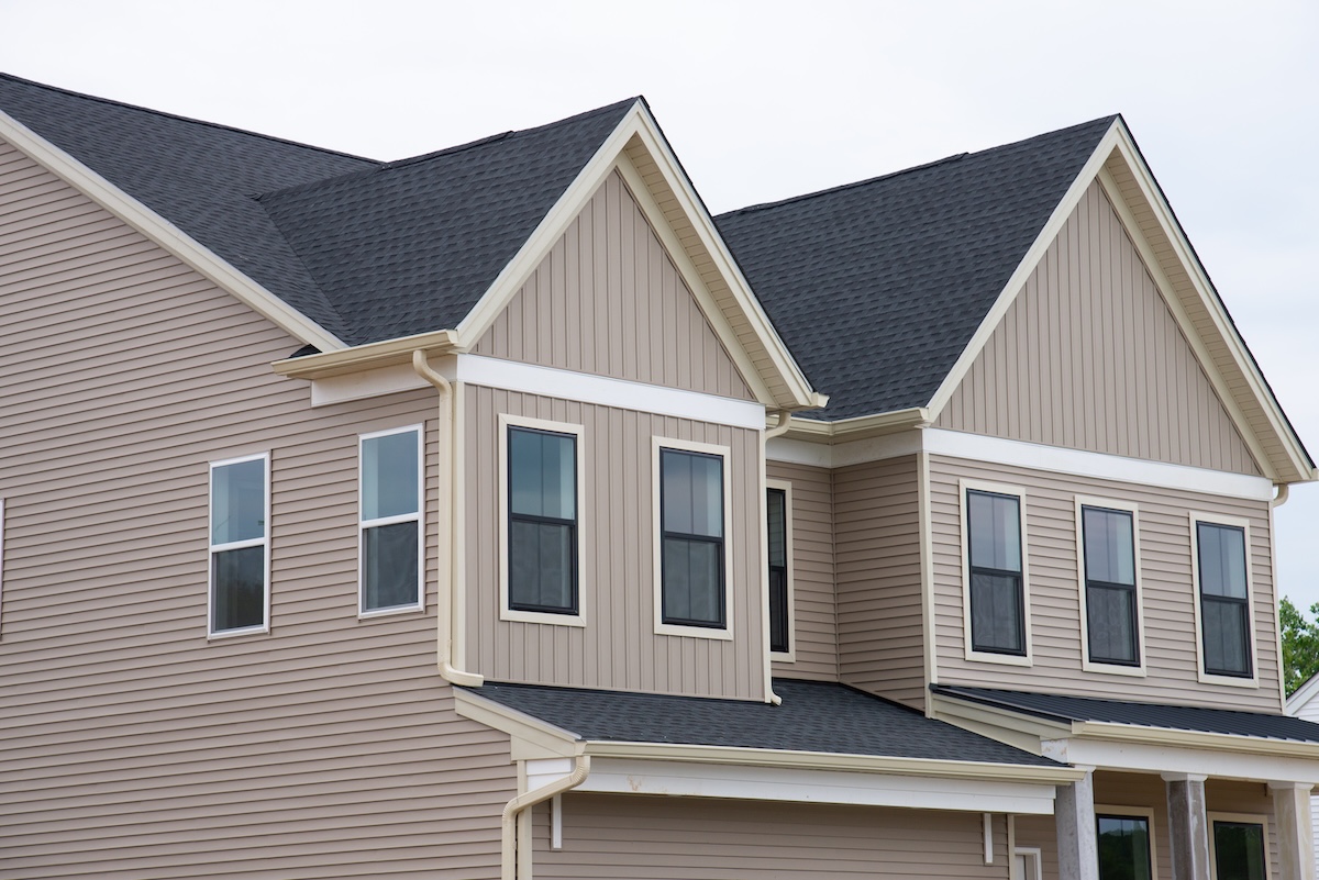 best asphalt shingles A close-up view of the exterior of a modern, two-story beige house with black-shingled roofs, white trim, multiple windows, and cloudy sky in the background.
