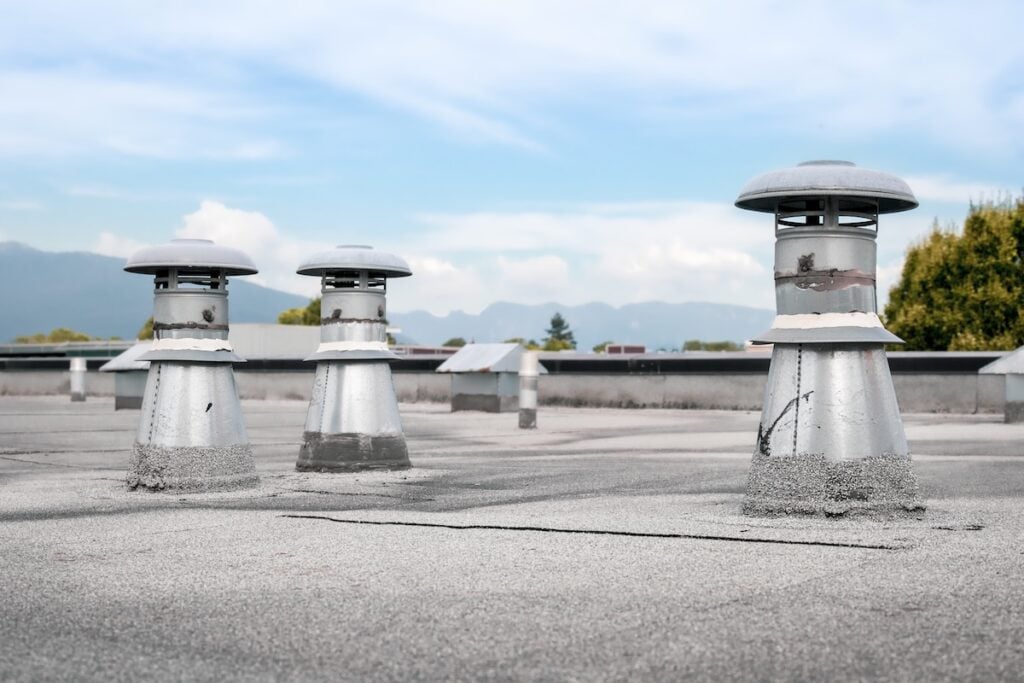 modified bitumen roof repair Three metal rooftop vents stand on a flat, gray rooftop under a blue sky with clouds. Trees and distant mountains are visible in the background.