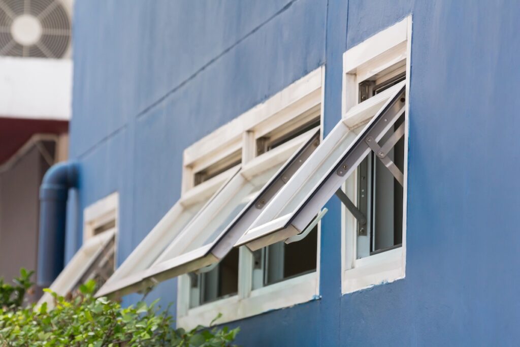 types of windows Three rectangular windows set in a blue wall, each open outward with metal frames. Green leaves are visible below, and part of an air conditioning unit is in the background.