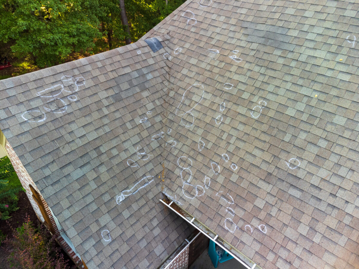 Aerial view of a house roof with numerous white chalk circles marking hail damage on the shingles, surrounded by trees.
