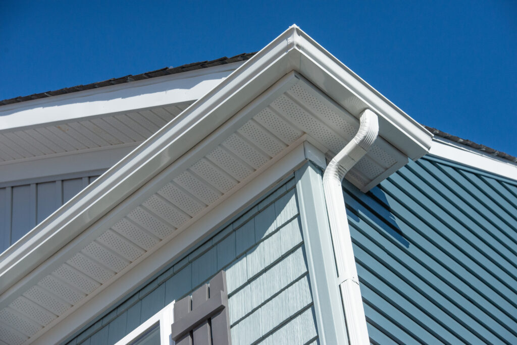 gutter installation cost Close-up view of a house exterior showing blue siding, white trim, a roof overhang, soffit, and a white rain gutter downspout against a clear blue sky.