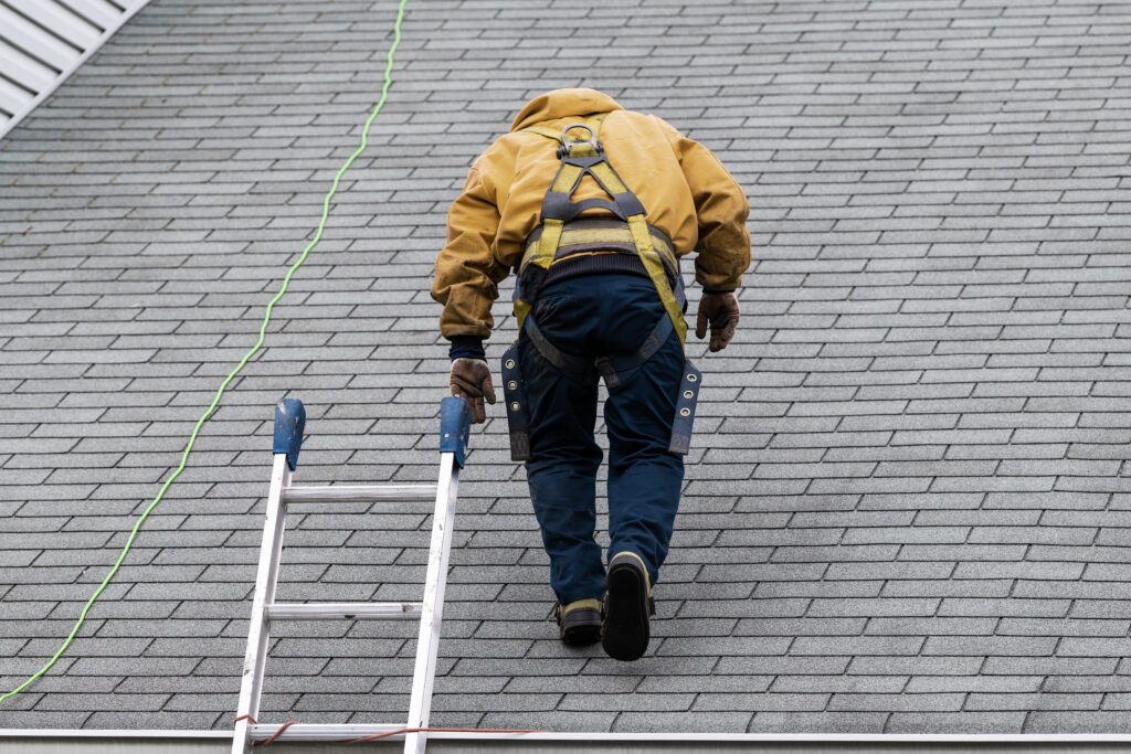 House during day with gray color Single Family Home and construction man in yellow uniform walking on roof shingles and ladder during repair