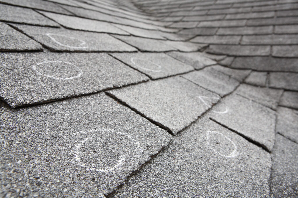 Close-up of gray asphalt roof shingles with several circular chalk marks indicating areas of damage or inspection points. The roof surface appears slightly uneven.