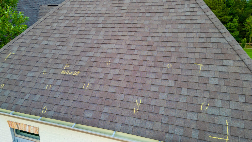 A roof with brown shingles has yellow chalk markings indicating areas for inspection or repair. Some markings include numbers and letters. Trees and grass are visible in the background.