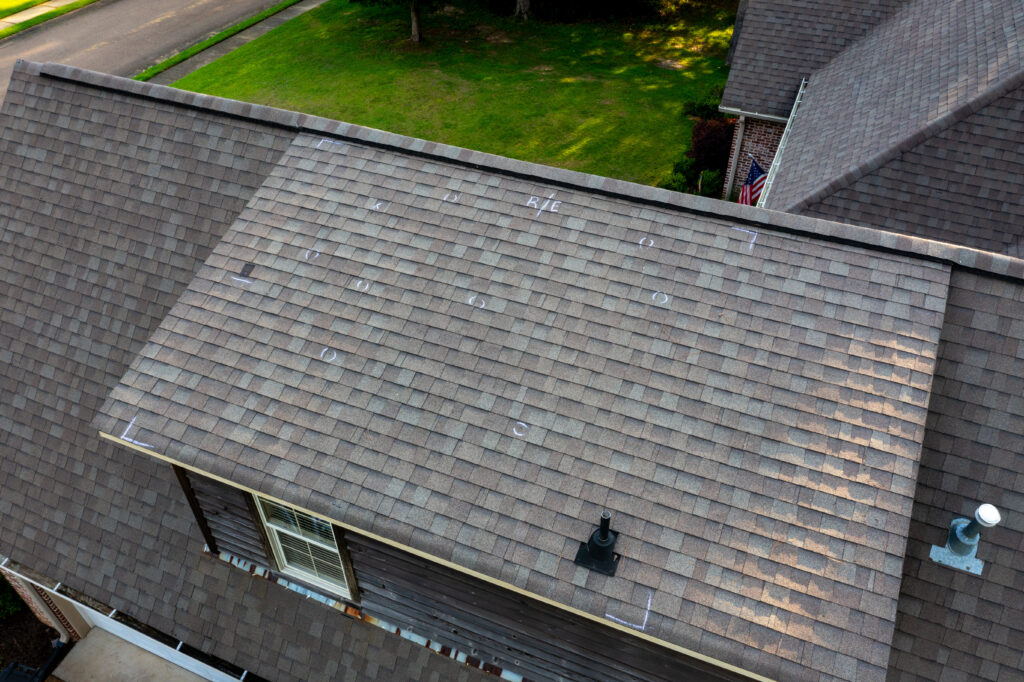 Aerial view of a house roof with visible chalk markings indicating inspection points, surrounded by green grass and neighboring rooftops. A vent and dormer window are also visible on the rooftop.