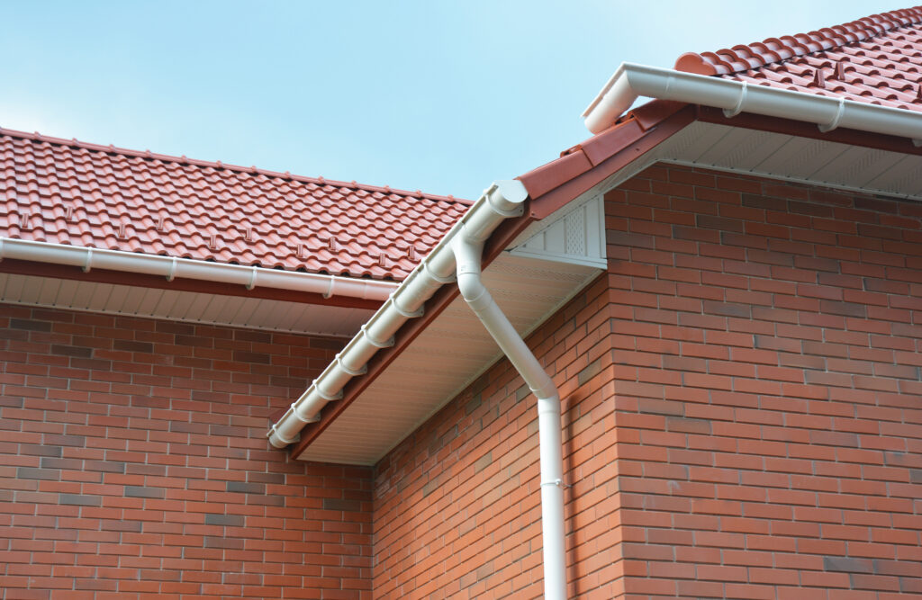 gutter installation cost Close-up view of the corner of a house with a red tiled roof, white rain gutters, and red brick walls under a clear blue sky.