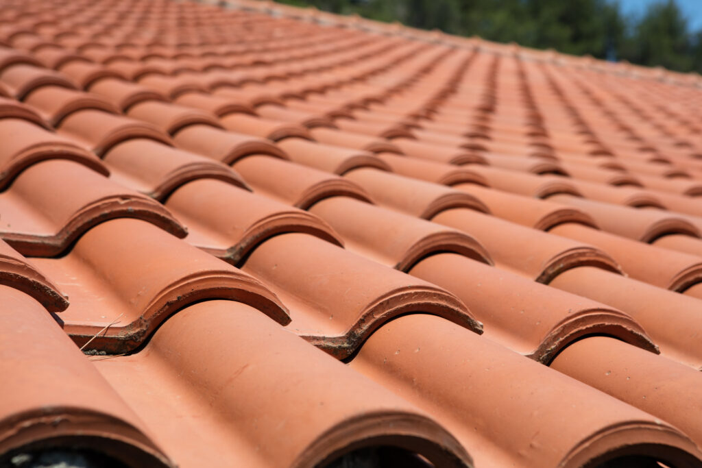 red tile roof Close-up view of a terracotta-tiled roof with rows of curved, reddish clay tiles, creating a repeating pattern. The tiles appear clean and well-maintained, with greenery visible in the blurry background.