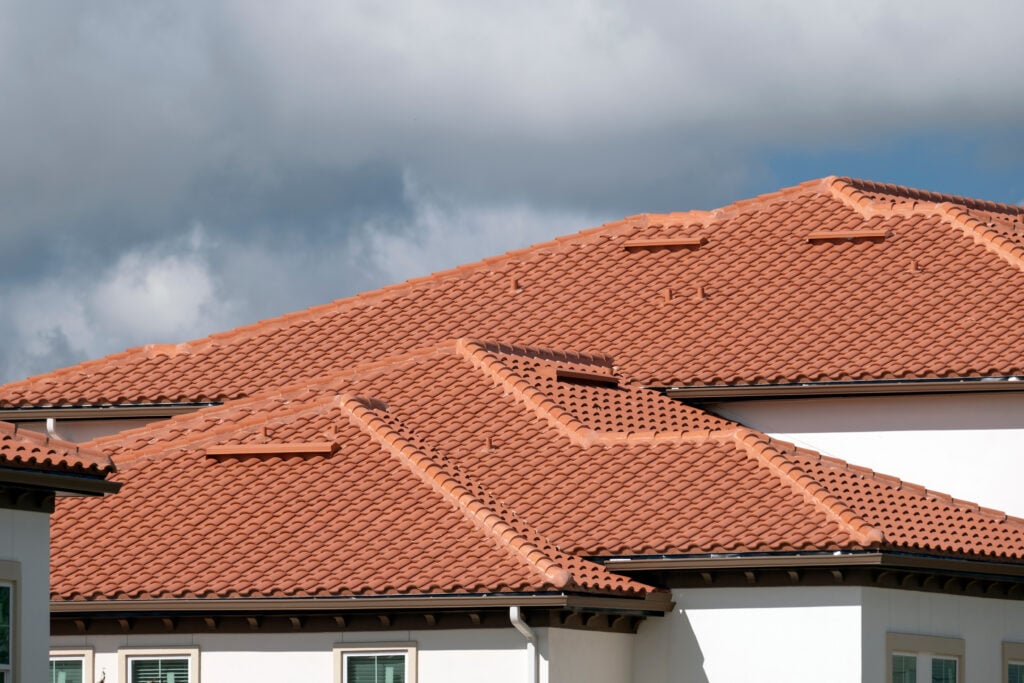 red tile roof Several houses with terracotta-colored clay tile roofs are shown under a partly cloudy sky. The roof tiles have a neat, uniform pattern and the house walls are white with a few windows visible at the bottom.