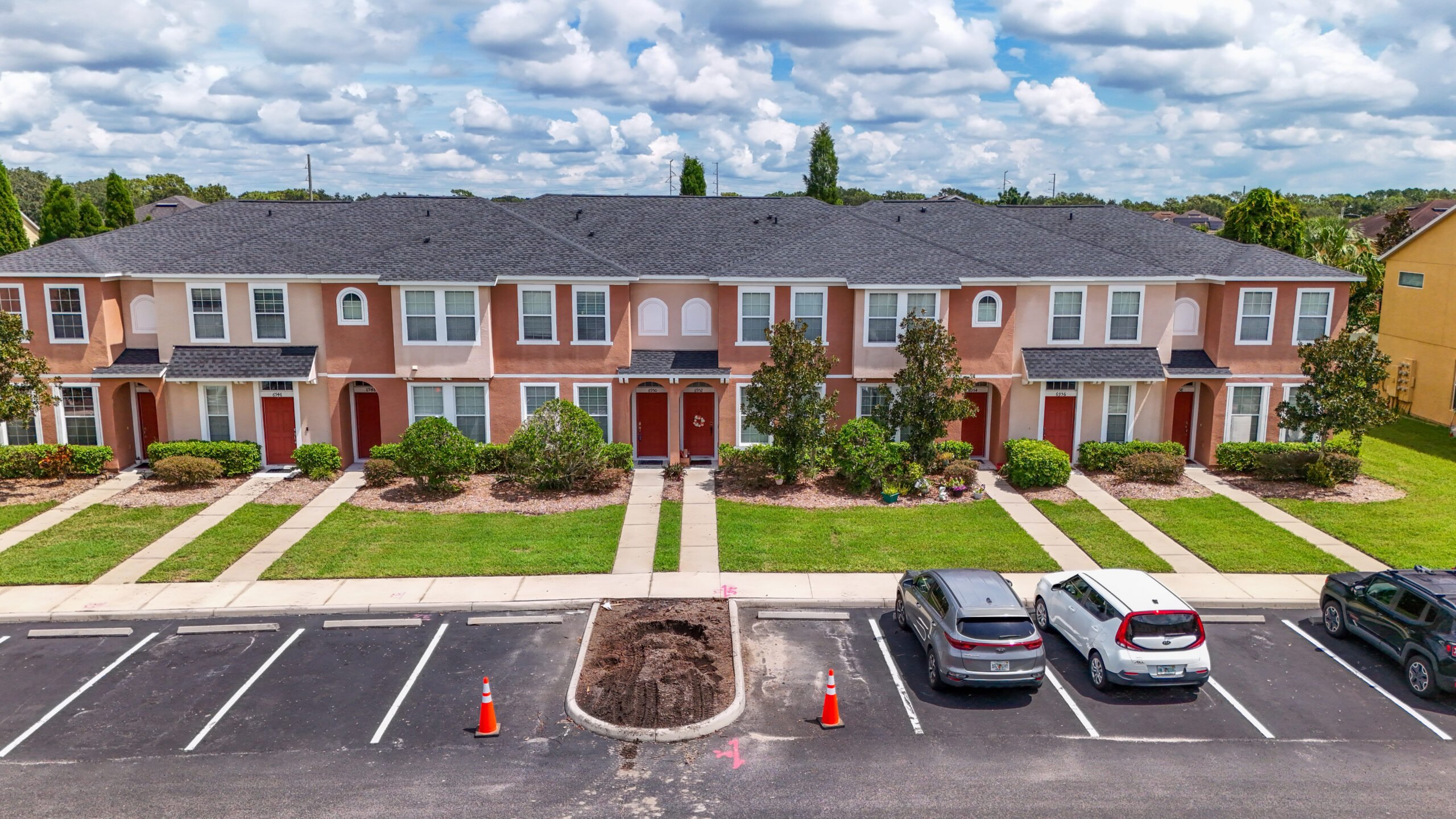 A row of two-story townhouses with peach and tan exteriors, green lawns, small trees, and bushes. Three cars are parked in front, and an empty parking space is marked with orange cones and dirt.