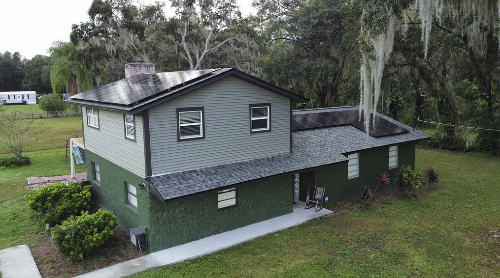 A two-story house with gray siding and a green lower level stands in a grassy yard. Solar panels, installed by a top roofing Tampa specialist, cover the roof, while trees with hanging moss surround the property. A mobile home is visible in the background.