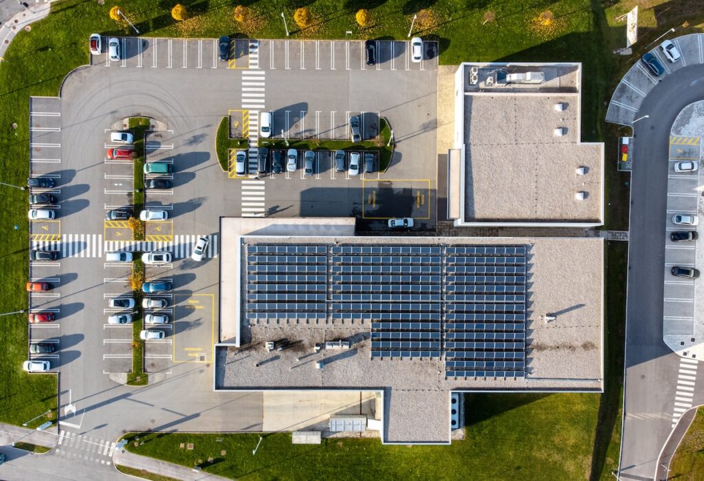 commercial roof inspection cost Aerial view of a building with solar panels on its roof, surrounded by parking lots with cars parked in rows, and green grassy areas with a few trees along the edges.