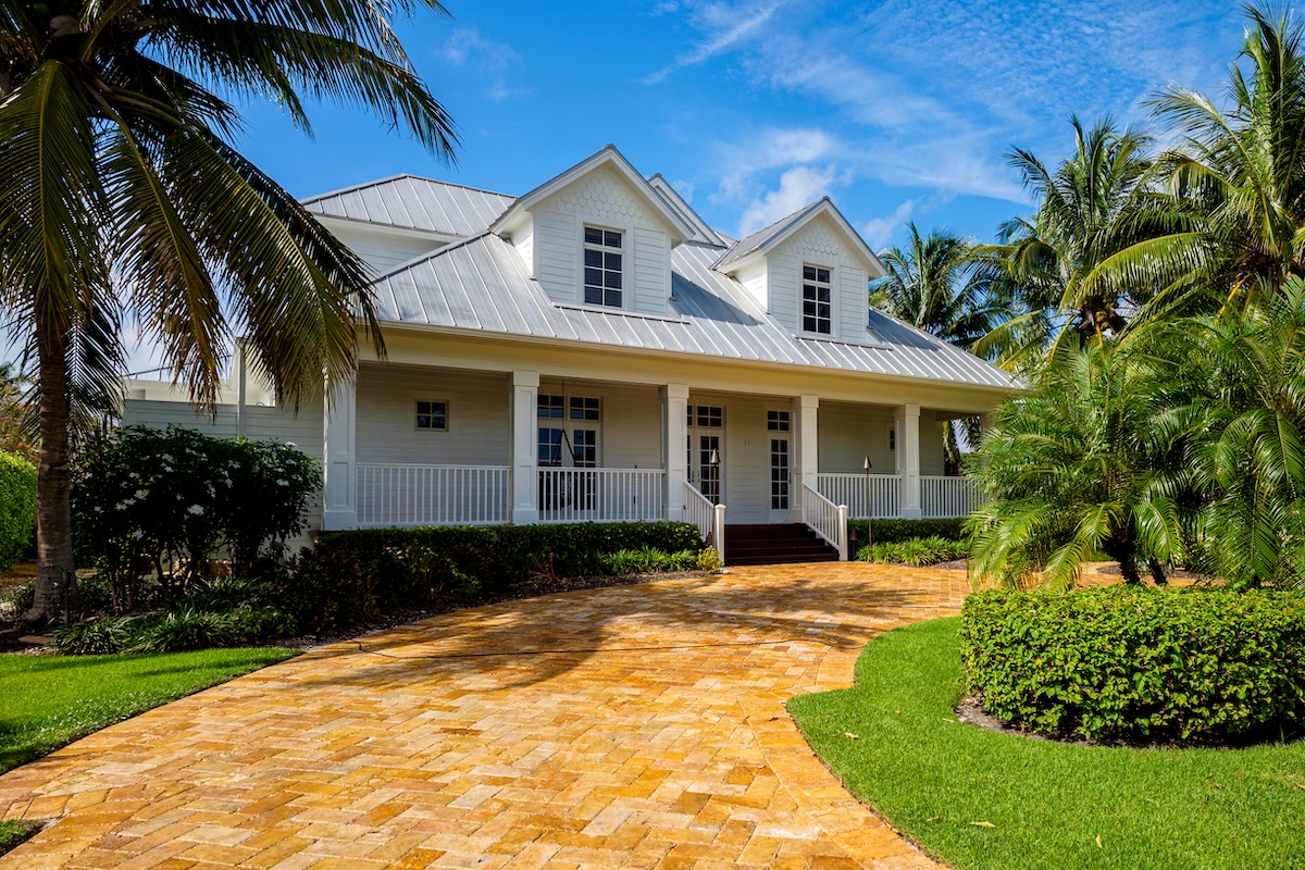 A white house with a metal roof and large front porch sits behind a curved brick driveway, surrounded by palm trees and lush landscaping—a perfect example of hurricane prep for roofs in Wesley Chapel under a bright blue sky.