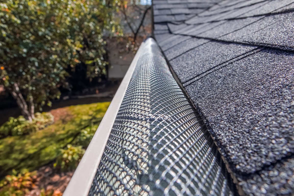 Close-up view of a house roof gutter covered with a metal mesh guard, demonstrating what is the best gutter guard for preventing debris from entering. Trees and lawn are visible in the blurred background.