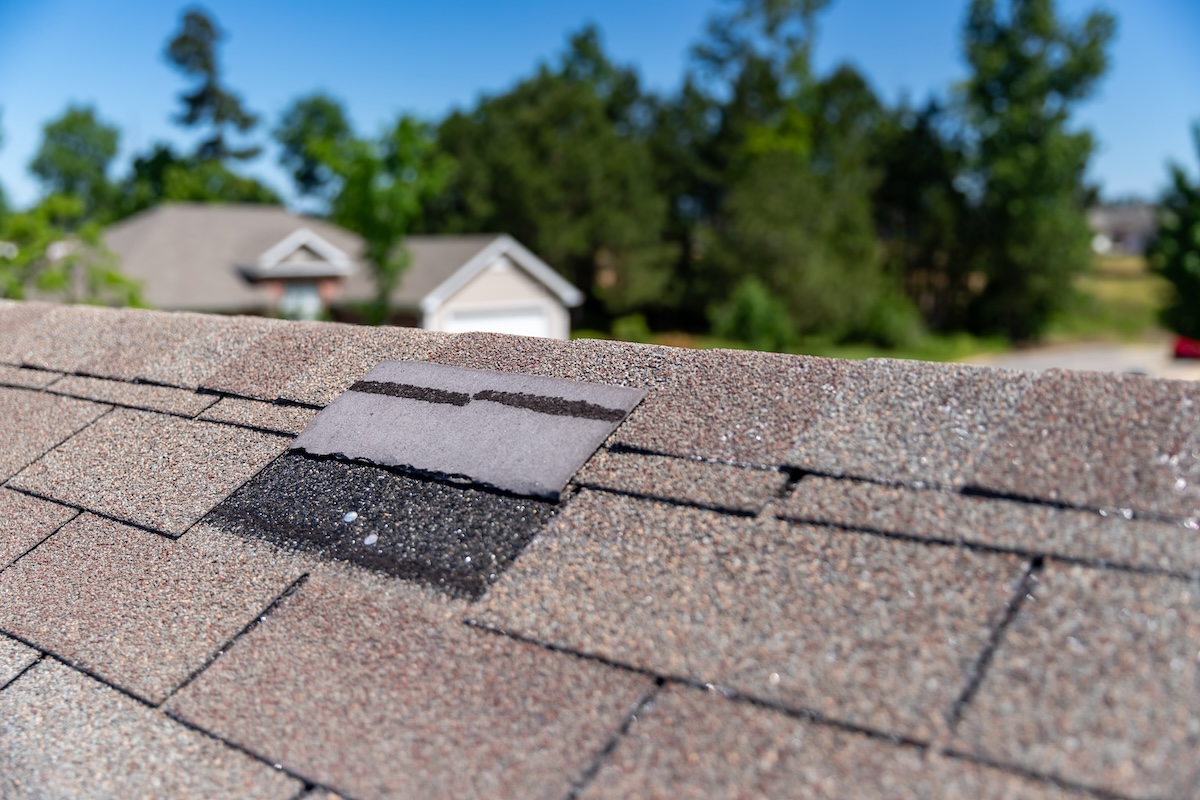 Close-up of a house roof with one missing asphalt shingle, exposing the dark underlayment beneath, with trees and houses blurred in the background on a sunny day.