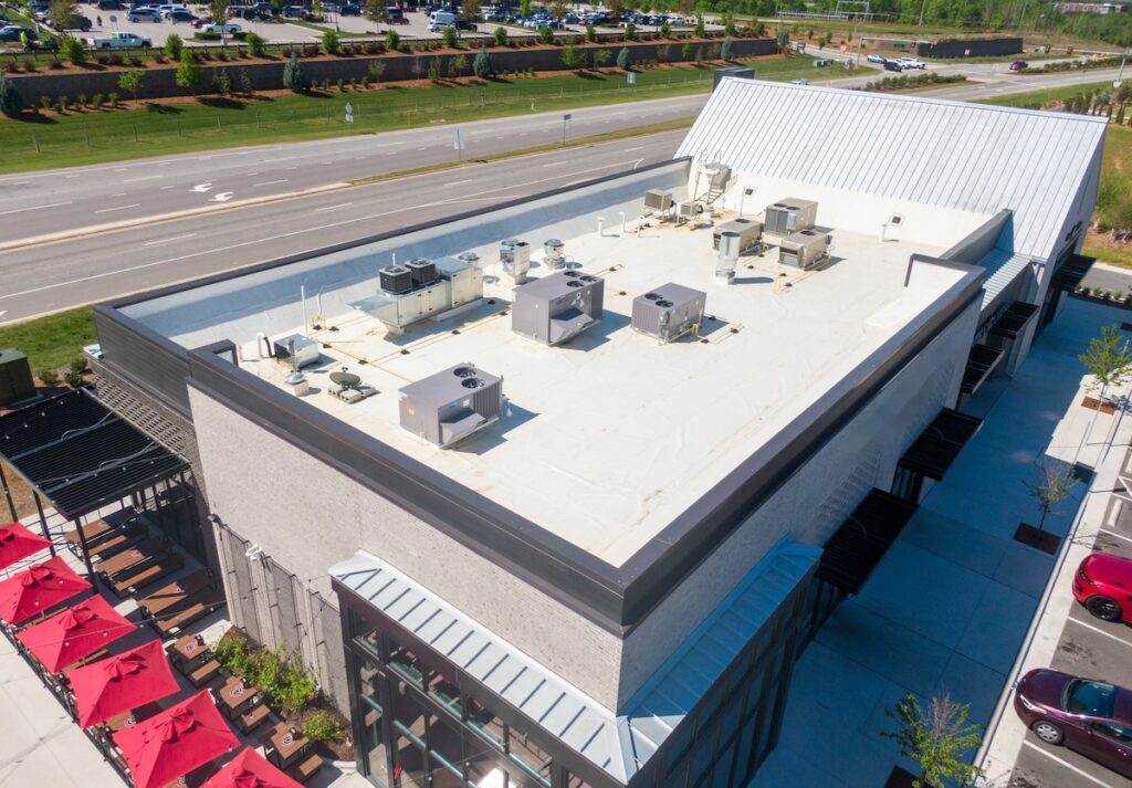 commercial roof inspection cost Aerial view of a commercial building with HVAC units on the white flat roof, located near a road and surrounded by parking lots and landscaping. Red umbrellas cover outdoor seating on one side.