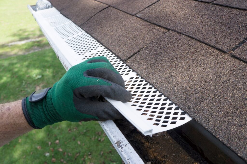 A person wearing a green glove installs a white plastic mesh gutter guard along the edge of a shingled roof, showing what is the best gutter guard for protecting gutters from debris.