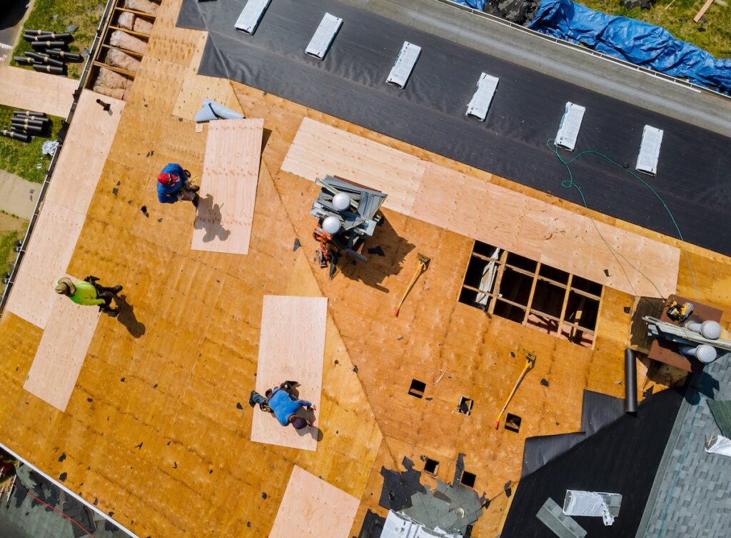 wesley chapel roof replacement cost Aerial view of four workers installing plywood sheets on a house roof under construction, with tools and materials scattered around the site—a typical scene when assessing Wesley Chapel roof replacement cost.