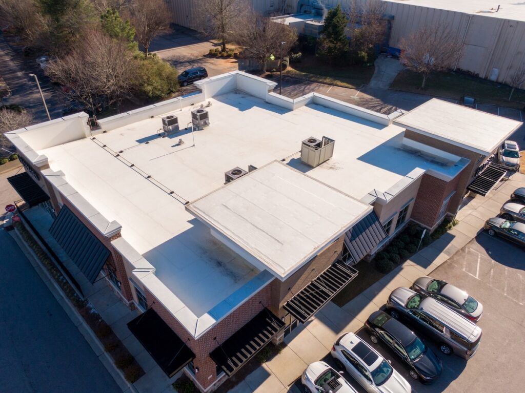 commercial roof inspection cost Aerial view of a commercial building with a white flat roof, HVAC units on top, brick walls, black awnings, and several parked cars in adjacent parking spaces. Trees and another building are visible in the background.
