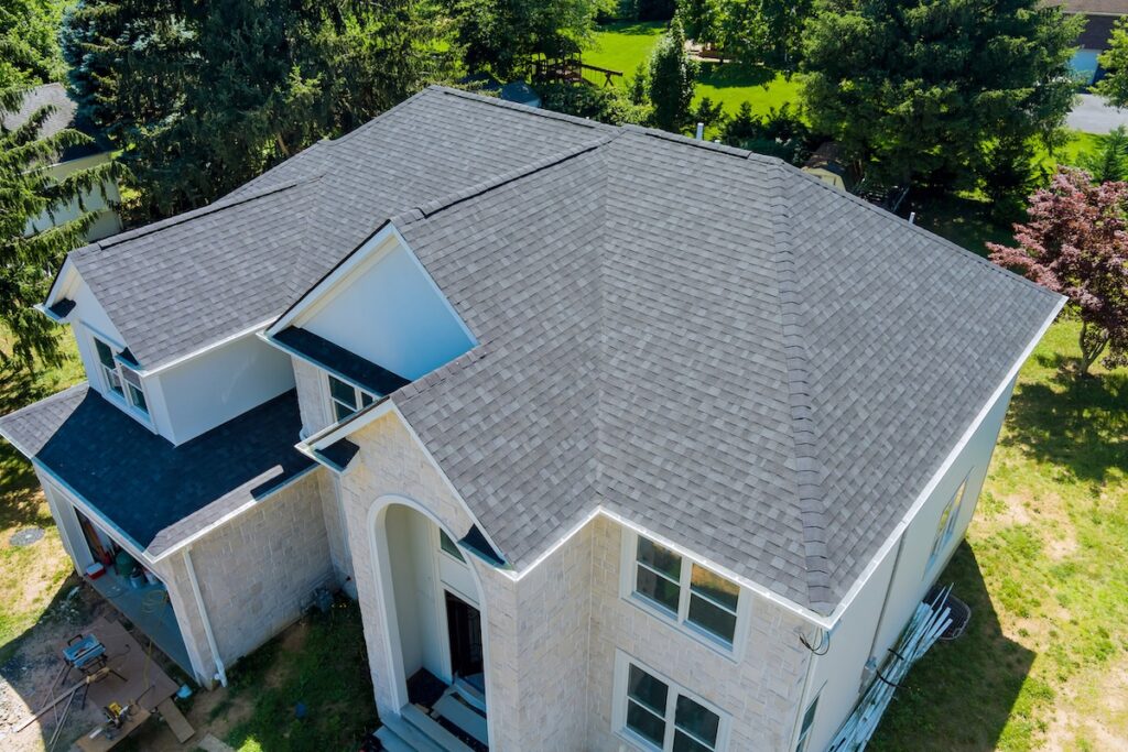 asphalt roof maintenance Aerial view of a large two-story house with light-colored stone walls and a gray shingle roof, surrounded by green trees and grass.
