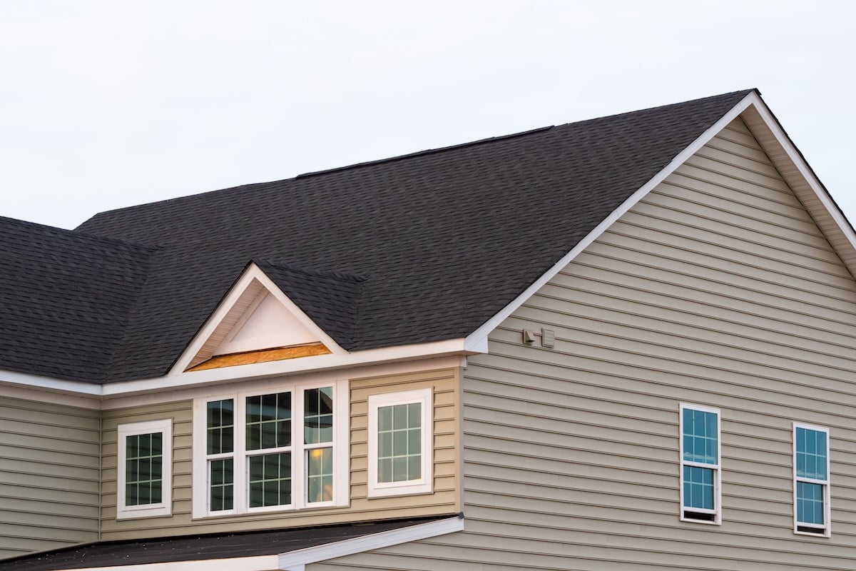 A close-up view of the upper portion of a beige house with dark gray shingles, several windows, and horizontal vinyl siding. The sky in the background is pale and clear.