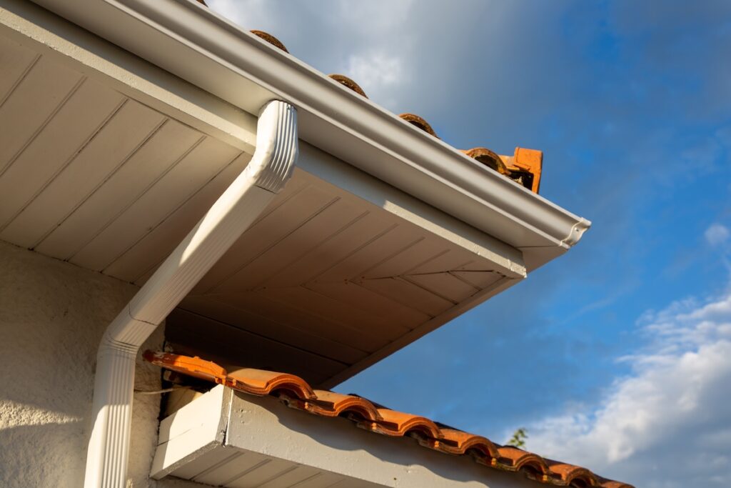 seamless gutters A close-up view of a house roof corner with orange tiles, a white seamless gutter, and a downspout against a partly cloudy blue sky.