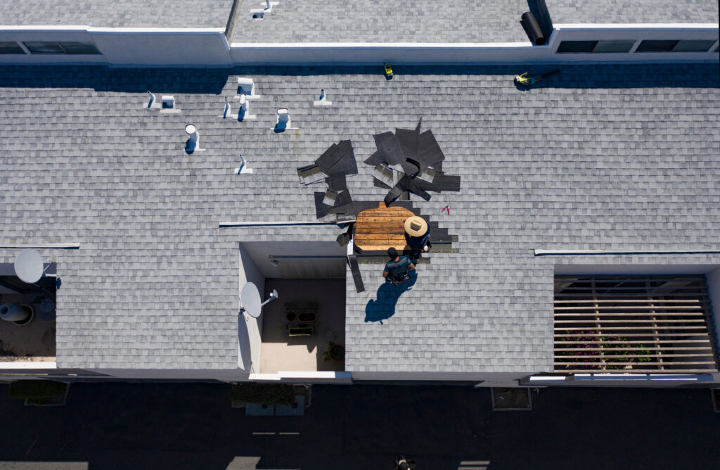 asphalt shingle roof leak repair An aerial view of a person working on the gray shingle roof of a building, with roofing materials scattered around. The worker wears a hat and is near a rectangular opening on the rooftop.