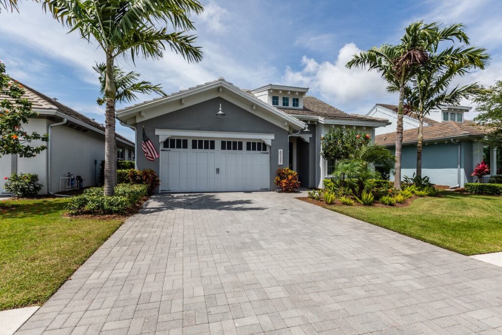 wesley chapel storm damage insurance A modern gray house with a two-car garage and an American flag by the entrance, surrounded by palm trees, green lawn, and landscaped gardens on a sunny day.