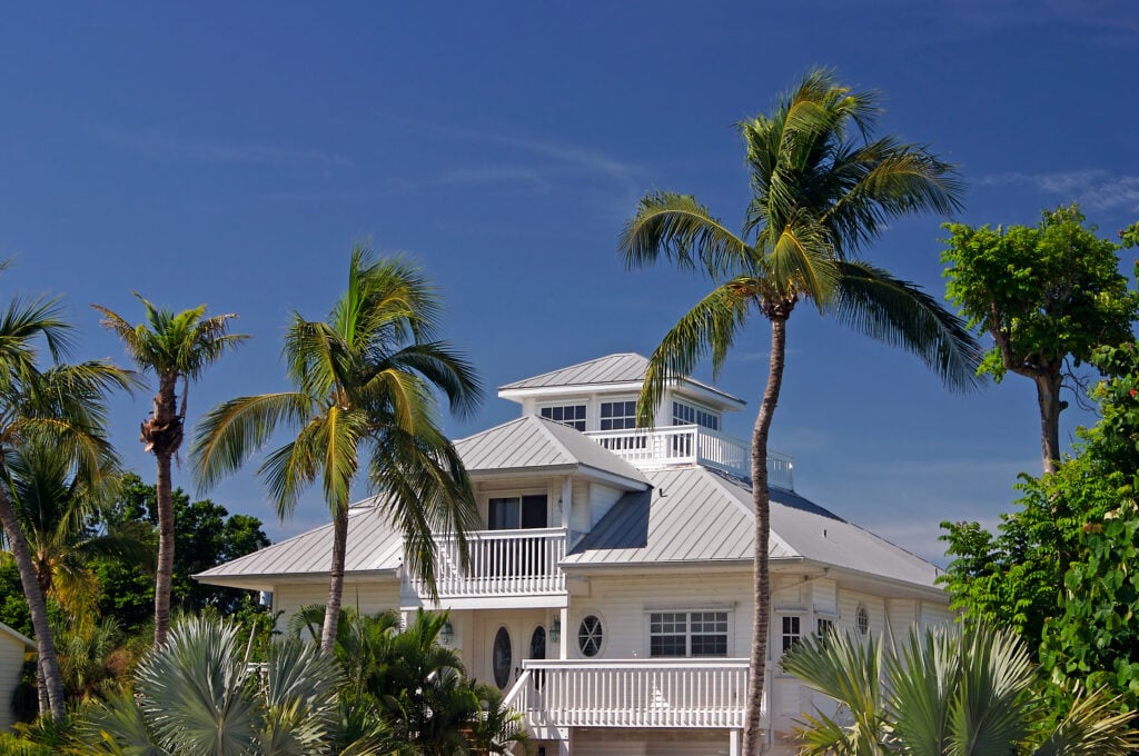 hurricane prep for roofs Wesley chapel A white, multi-story beach house with a metal roof and balconies is surrounded by tall palm trees and lush greenery under a clear blue sky, showcasing the importance of hurricane prep for roofs in Wesley Chapel.