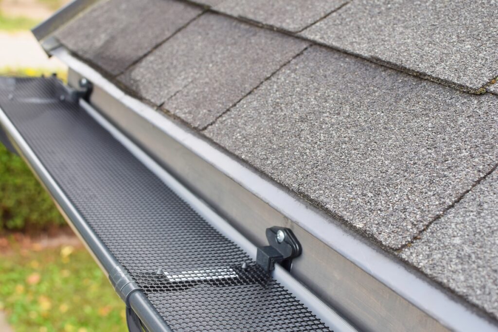 Close-up view of a house roof with asphalt shingles and a metal mesh gutter guard installed to prevent debris from entering the rain gutter, showcasing what is the best gutter guard for effective protection.