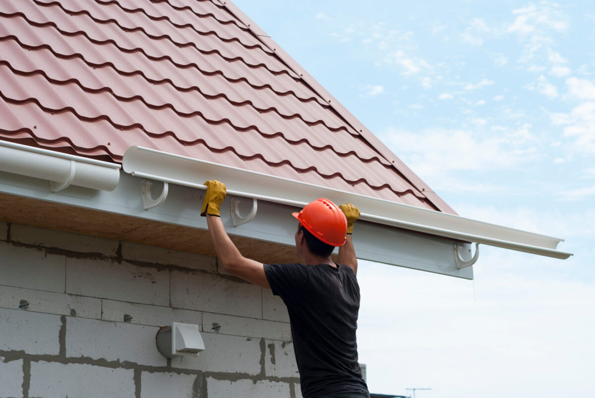 A person wearing a hard hat and yellow gloves installs seamless gutters along the edge of a red metal roof on a brick house, under a partly cloudy sky.