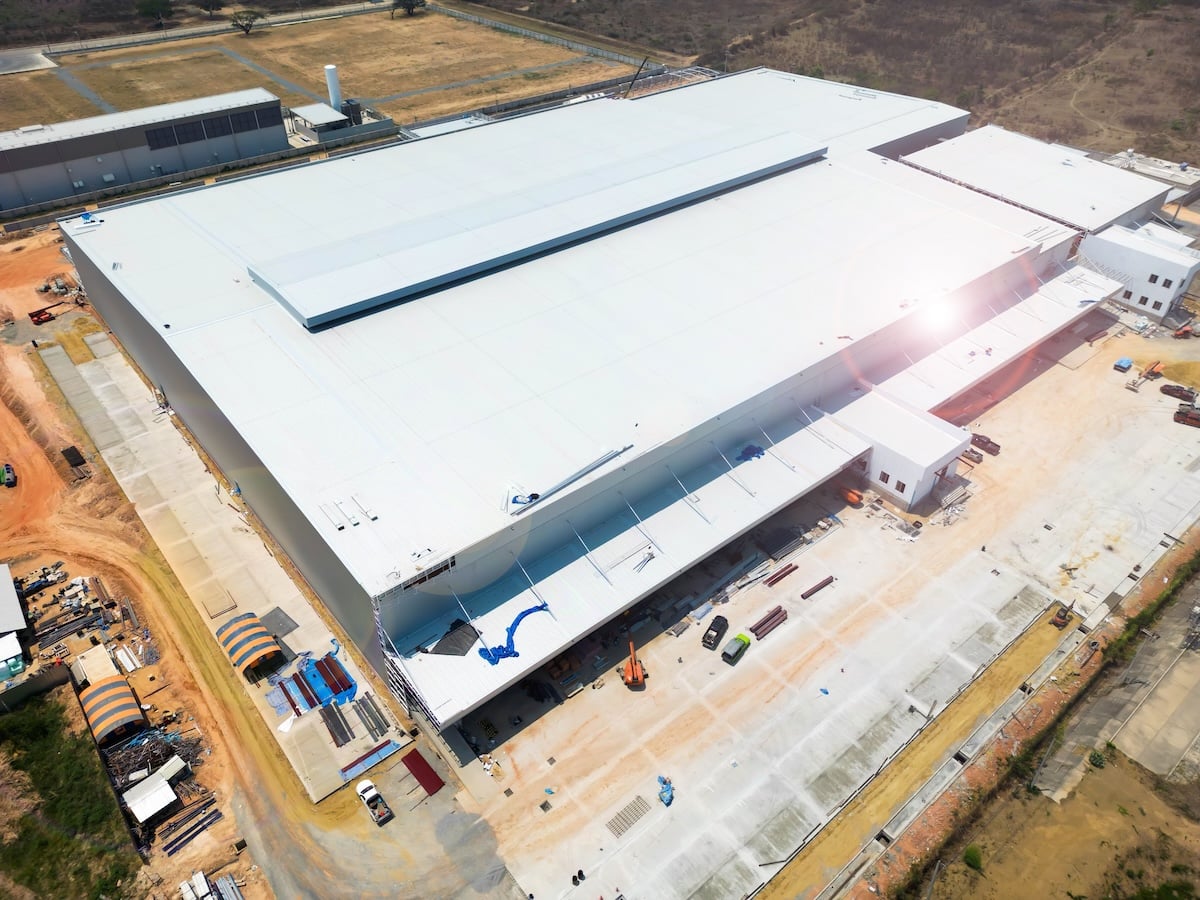 Aerial view of a large, modern industrial warehouse with a white roof, surrounded by paved areas, construction materials, and vehicles on a mostly barren landscape.