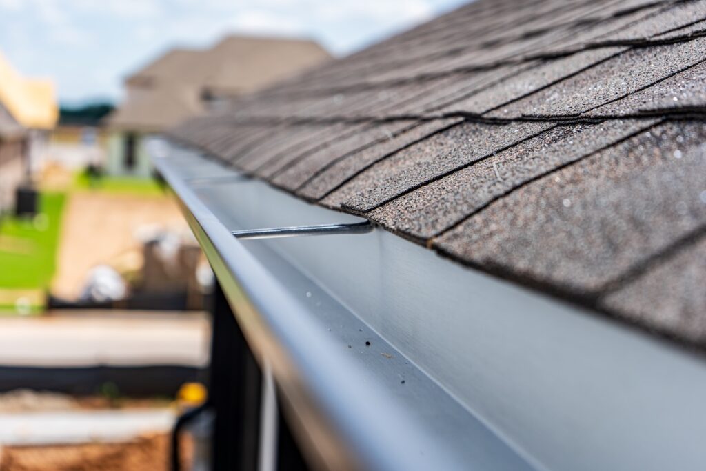 seamless gutters Close-up view of a house roof with brown asphalt shingles and a clean, new seamless gutters system installed along the edge, set against a blurred background of other houses and a sunny outdoor scene.
