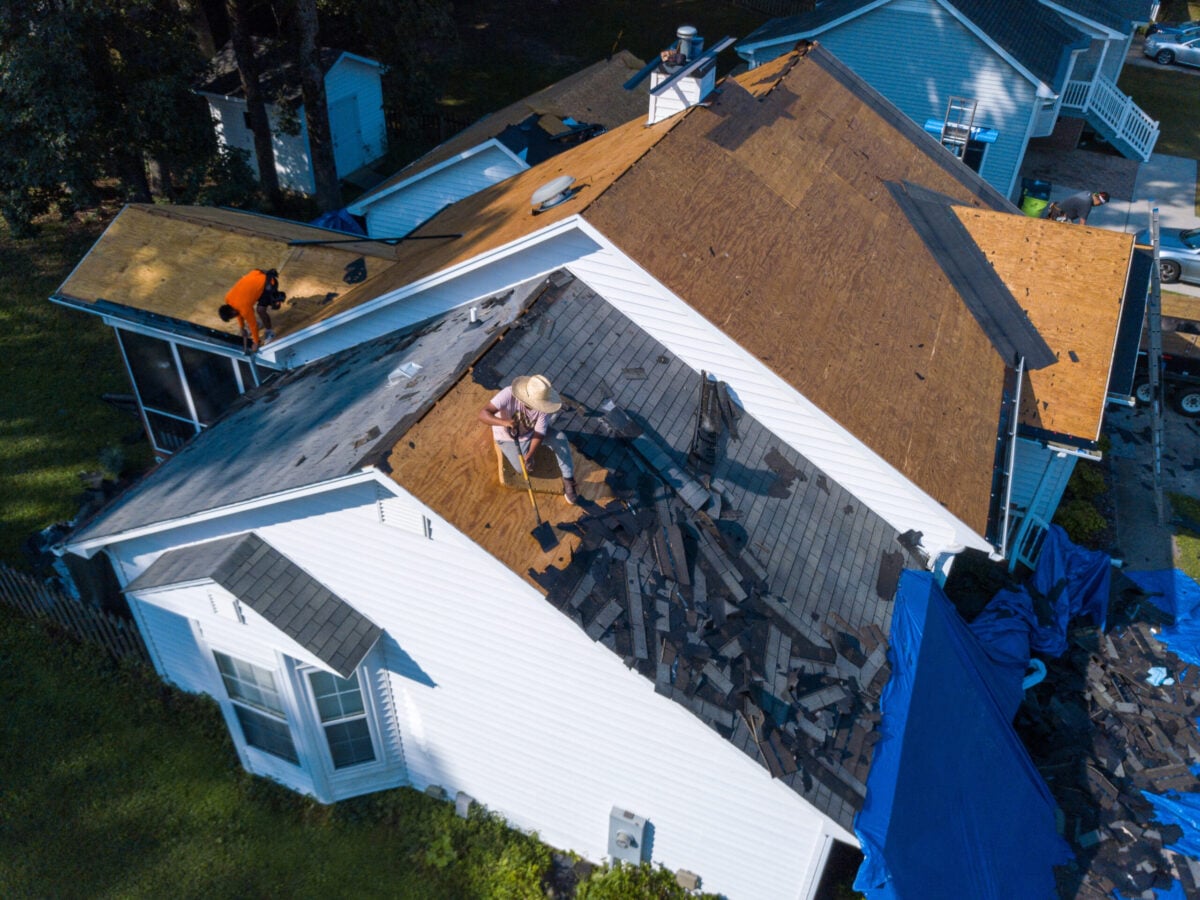 A person wearing a hat works on removing old shingles from the roof of a white house, while another tackles a nearby roof. Roofing materials and debris are scattered, with blue tarps below—typical during a Wesley Chapel roof replacement cost estimate.