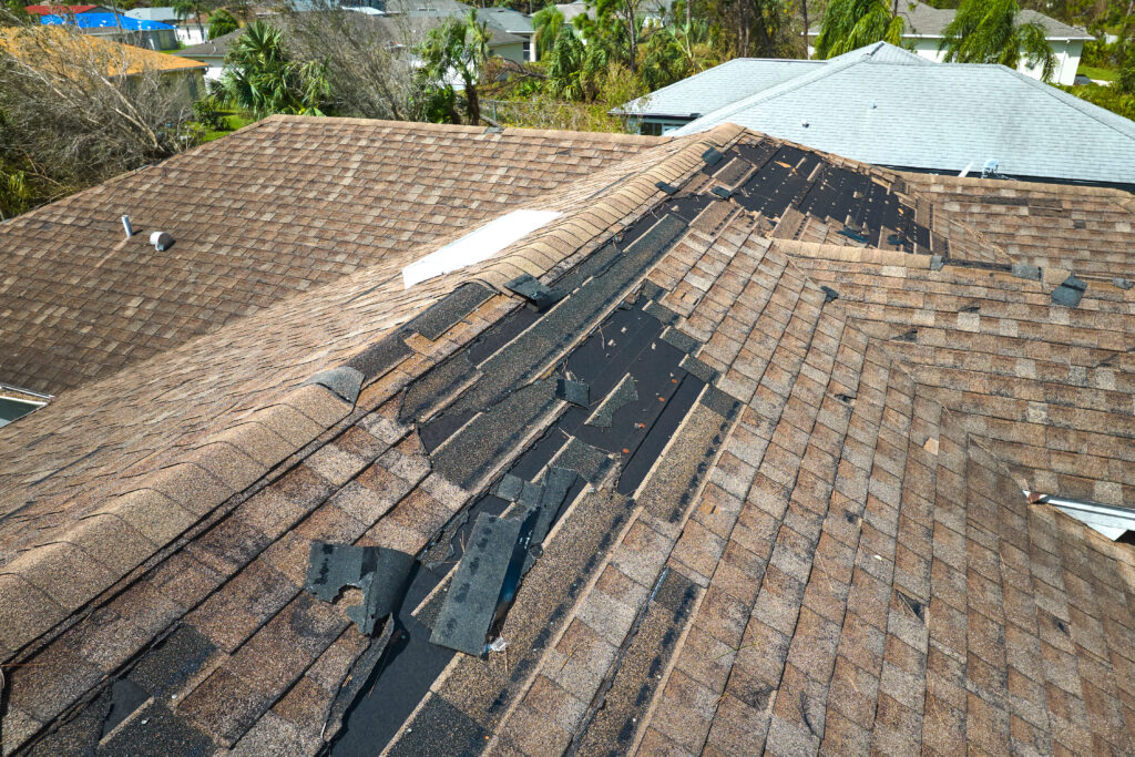 wesley chapel storm damage insurance A house roof with damaged and missing shingles, exposing black underlayment beneath. The damage runs along the ridge, while other shingles remain intact. Nearby rooftops and trees are visible in the background.