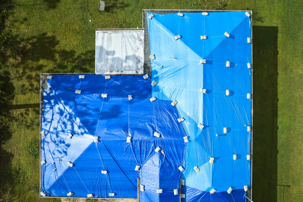 Aerial view of a house roof covered with large blue tarps and sandbags—classic hurricane prep for roofs in Wesley Chapel—surrounded by lush green grass and trees.