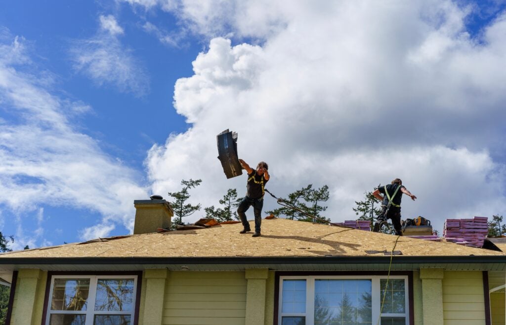 wesley chapel roof replacement cost Two workers are on the roof of a house—one lifting shingles, the other near stacks of materials. With trees and scattered clouds in view, this scene highlights a typical day on a job where homeowners consider Wesley Chapel roof replacement cost.