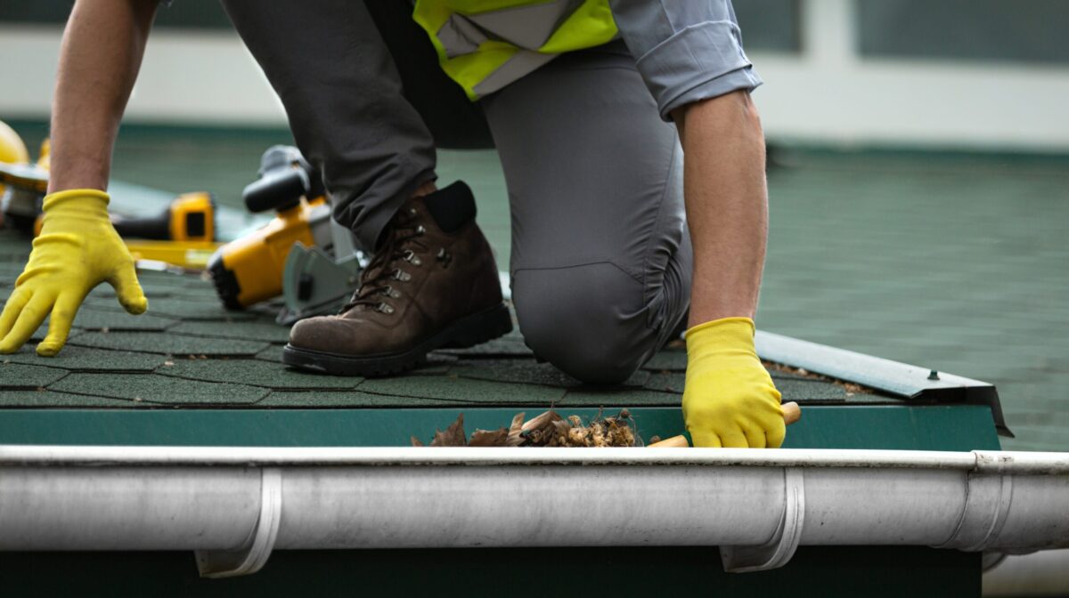 A person wearing yellow gloves and work boots is cleaning leaves and debris from a house gutter while kneeling on a green shingle roof. Tools are visible in the background.