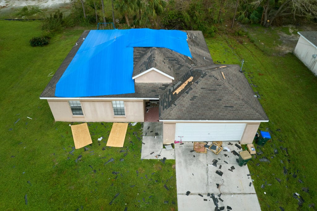 A tan house with roof damage, a large blue tarp covering part of the roof, shingles scattered on the yard and driveway, and plywood leaning against the front wall—a common sight when considering Wesley Chapel roof replacement cost.