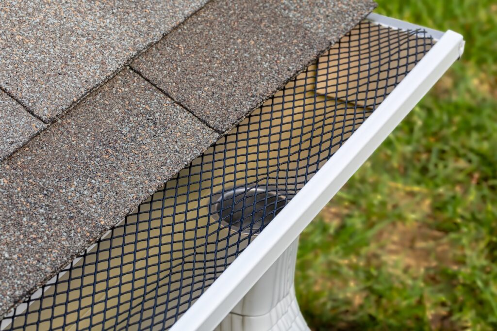 Close-up of a house roof edge with shingles, a mesh gutter guard covering the rain gutter, and a downspout, showing why many ask, "what is the best gutter guard?" Green grass is visible in the background.