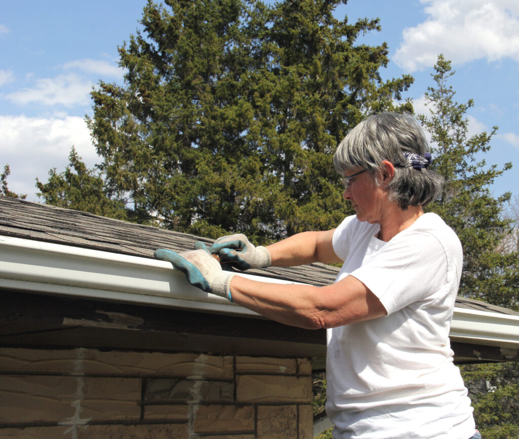 gutter cleaning A woman wearing gloves and a white shirt cleans debris from the gutter of a house, standing on a ladder beside a roof, with trees and a blue sky in the background.