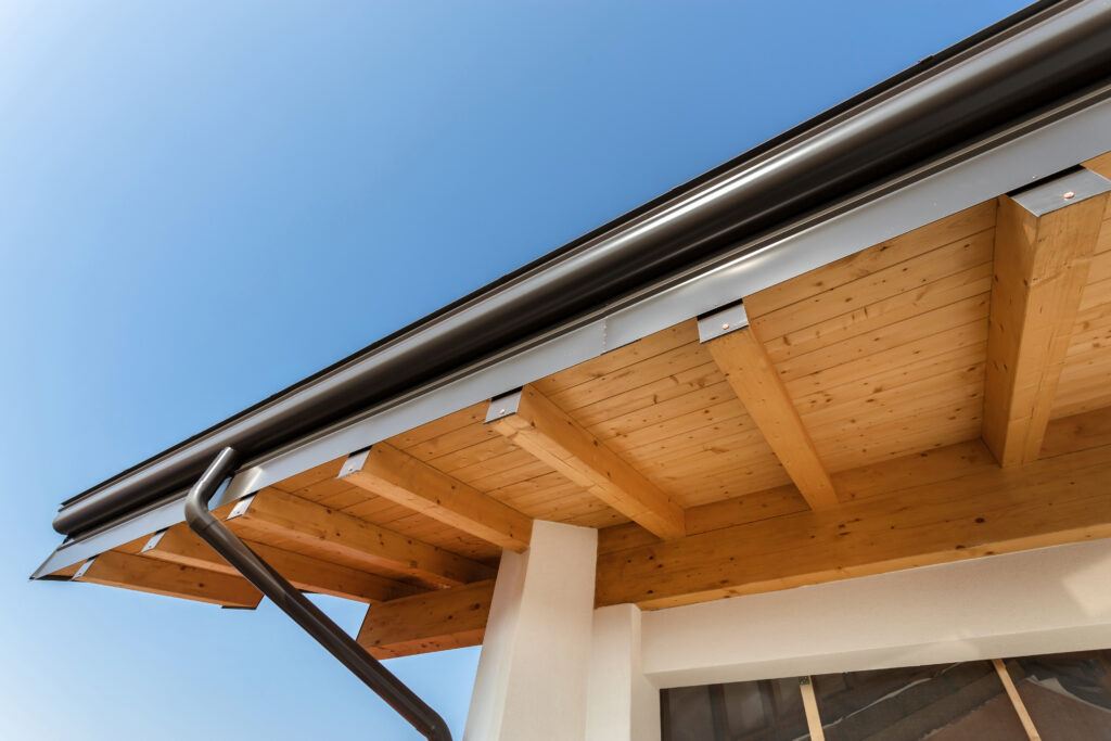 seamless gutters Close-up view of a house’s wooden roof overhang with exposed beams, seamless gutters, and a black rain gutter set against a blue sky in the background.